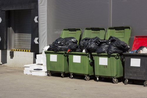 A row of four green wheelie bins with partially open lids, filled with black rubbish bags, are positioned along a concrete surface outside a commercial building in Shepherds Bush. The bins are on caster wheels, and the black rubbish bags appear tightly packed and stacked up to the brim. To the right, a black skip container is visible, also filled with waste. In the background, part of a grey metallic roller shutter door and a section of a grey building façade are seen, along with a small white object, possibly debris or packaging material, on the ground nearby. The scene depicts typical waste disposal activity associated with commercial rubbish removal services in Shepherds Bush, highlighting waste collection and management at a business premises.