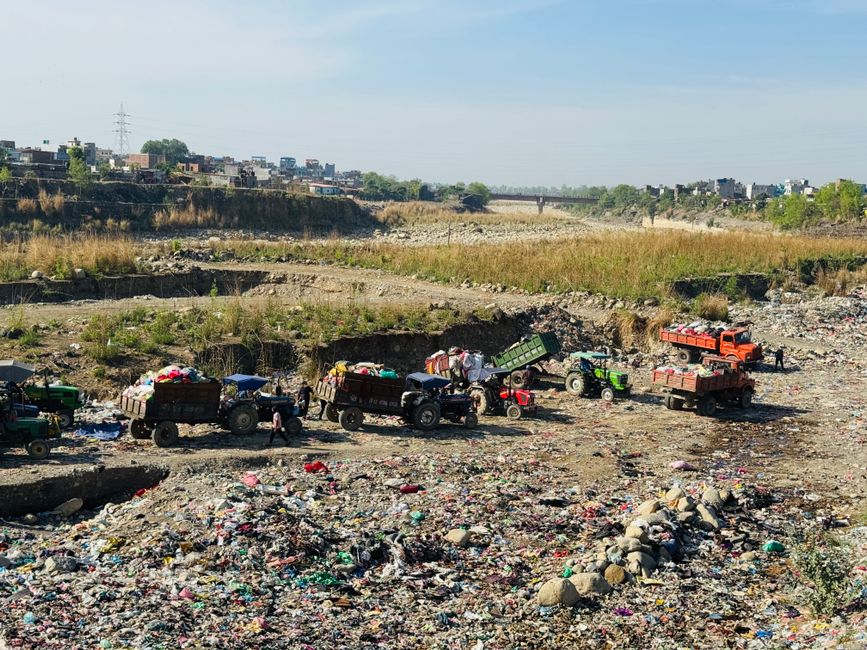 The image depicts an open, dry landfill site with several small tractors and trailers, some filled with mixed waste materials including textiles, plastics, and household trash. The vehicles are positi